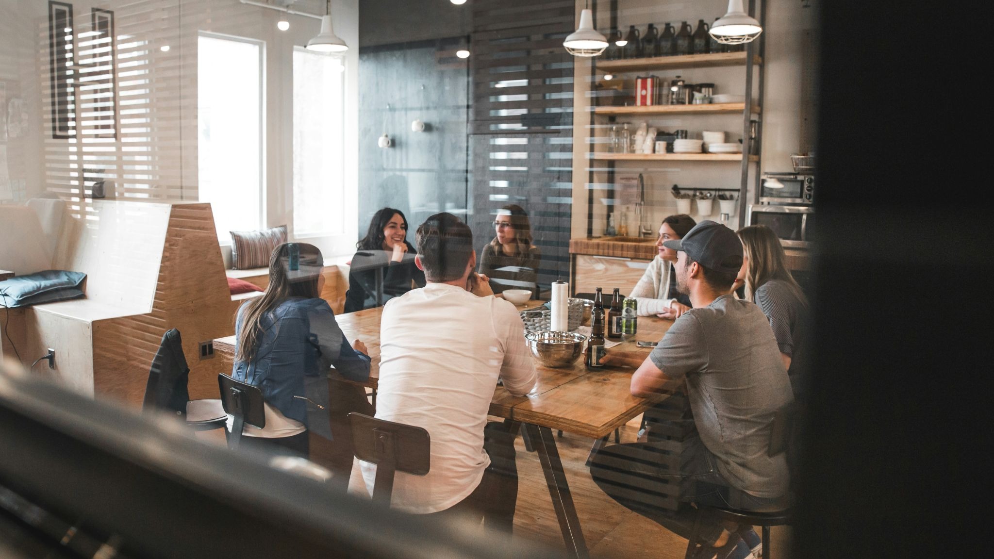 A group of colleagues chatting around a large wooden table in a bright, casual café-style workspace, viewed through a glass wall.